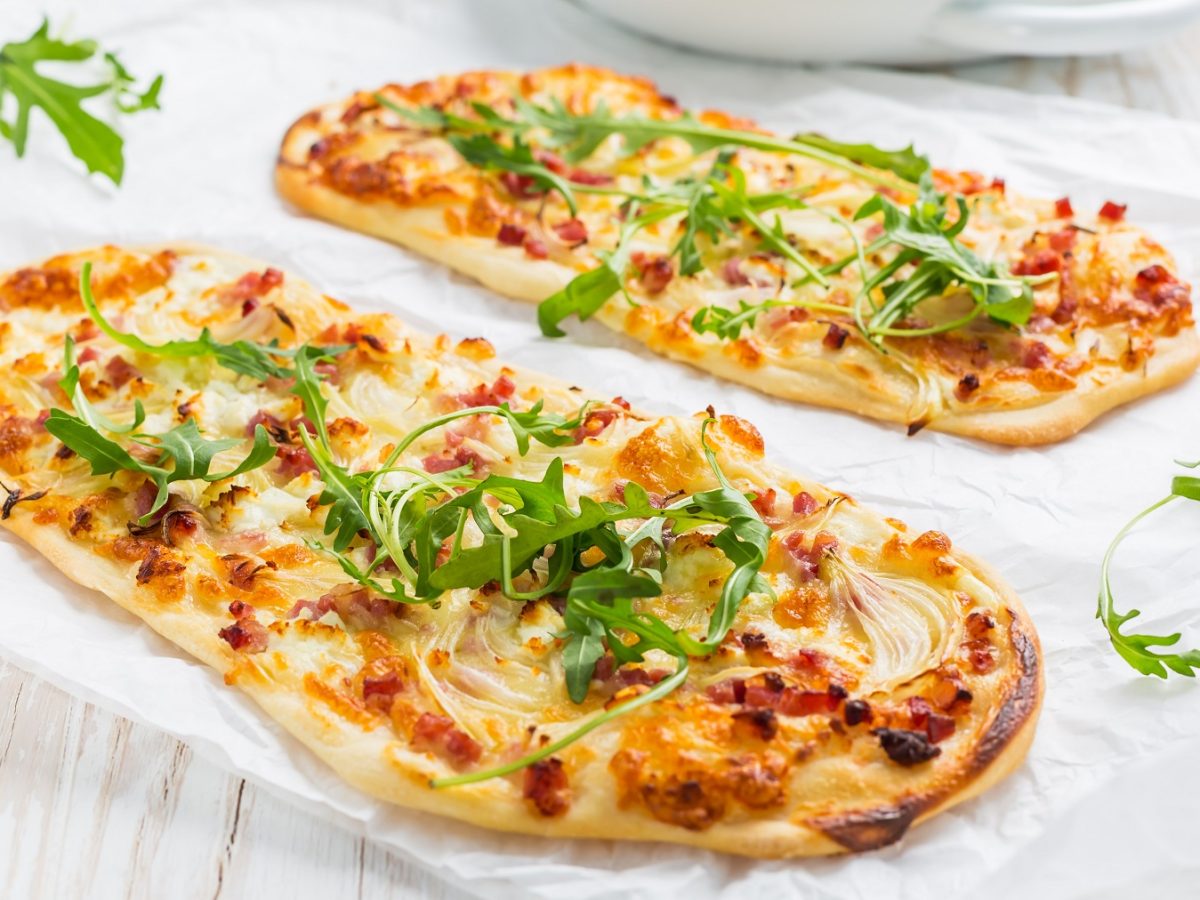 Zwei Flammkuchen auf einem weißen Tisch, mit Rucola bestreut von der Seite fotografiert.