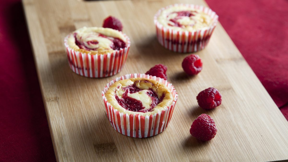 Drei Käsekuchen-Muffins mit frischen Himbeeren auf einem hellen Schneidebrett und rotem Untergrund. Alles von schräg oben fotografiert.