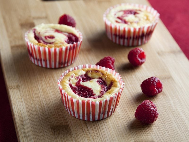 Drei Käsekuchen-Muffins mit frischen Himbeeren auf einem hellen Schneidebrett und rotem Untergrund. Alles von schräg oben fotografiert.