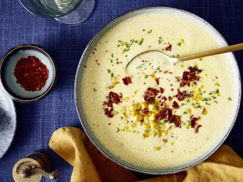 Mais-Cremesuppe in einer grauen Schüssel auf dunkelblauem Hintergrund. Links davon ein Schälchen mit Chili und unten auf dem Bild ein gelbes Geschirrtuch.