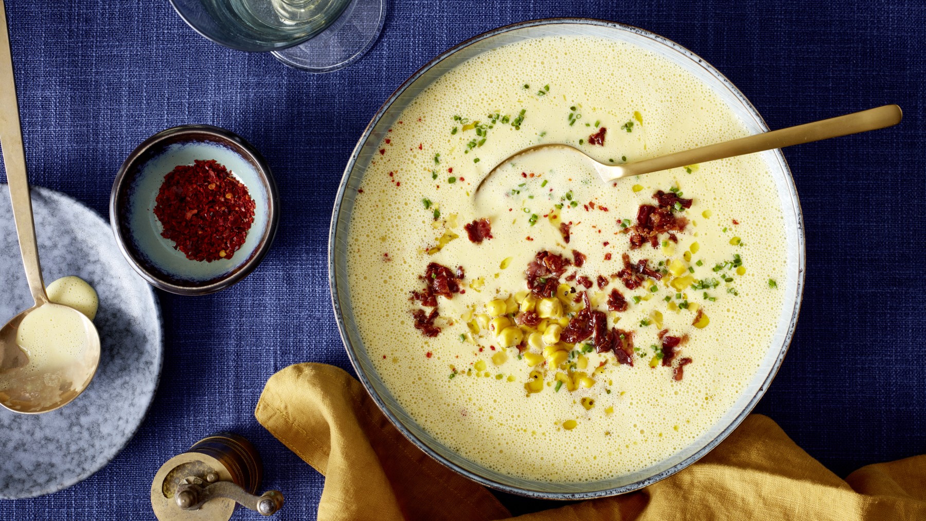 Mais-Cremesuppe in einer grauen Schüssel auf dunkelblauem Hintergrund. Links davon ein Schälchen mit Chili und unten auf dem Bild ein gelbes Geschirrtuch.