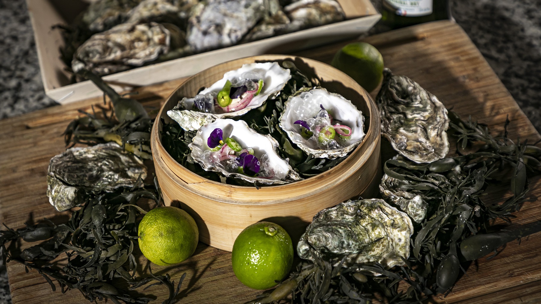 Gedämpfte Astern mit Toppings in einem Korb, daneben frische Austern und Limetten schräg von oben fotografiert.