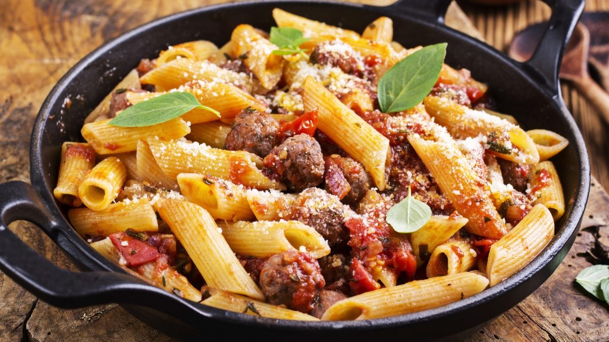 Eine schwarze gusseiserne Pfanne mit der Salsiccia-Pasta auf einem rustikalen Holztisch nah und von der Seite fotografiert.