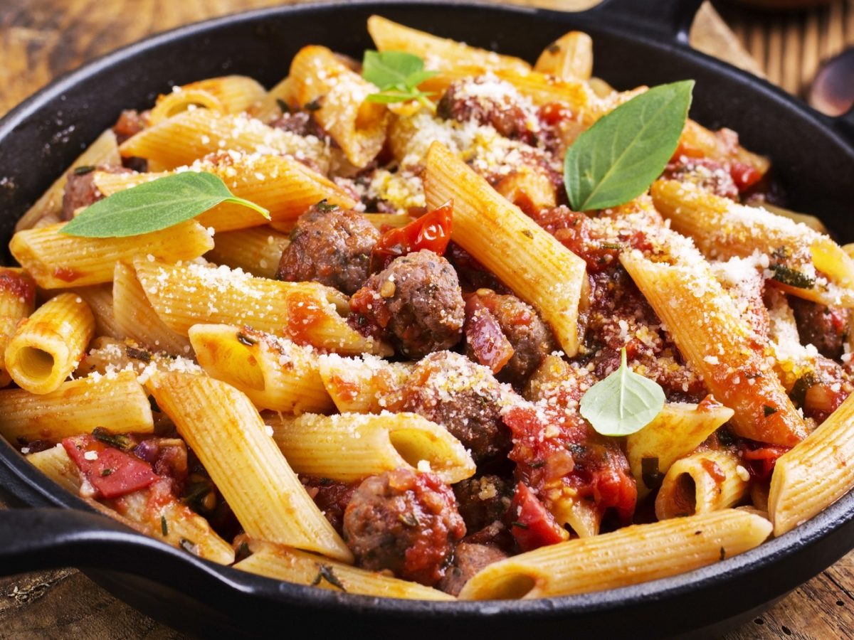 Eine schwarze gusseiserne Pfanne mit der Salsiccia-Pasta auf einem rustikalen Holztisch nah und von der Seite fotografiert.