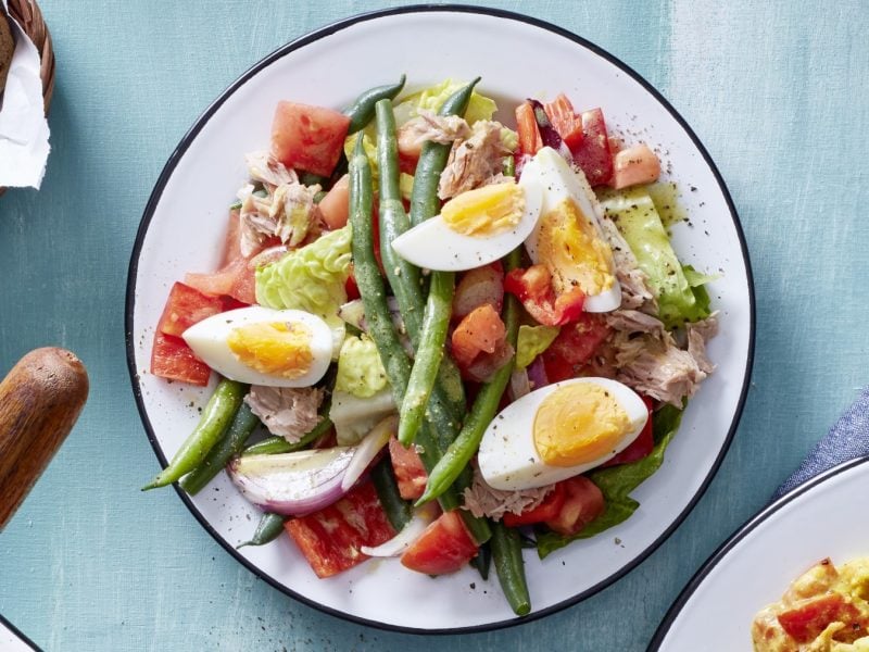 Salat mit Thunfisch und Ei in einer weißen Schüssel auf hellblauem Hintergrund. Links davon etwas Brot.