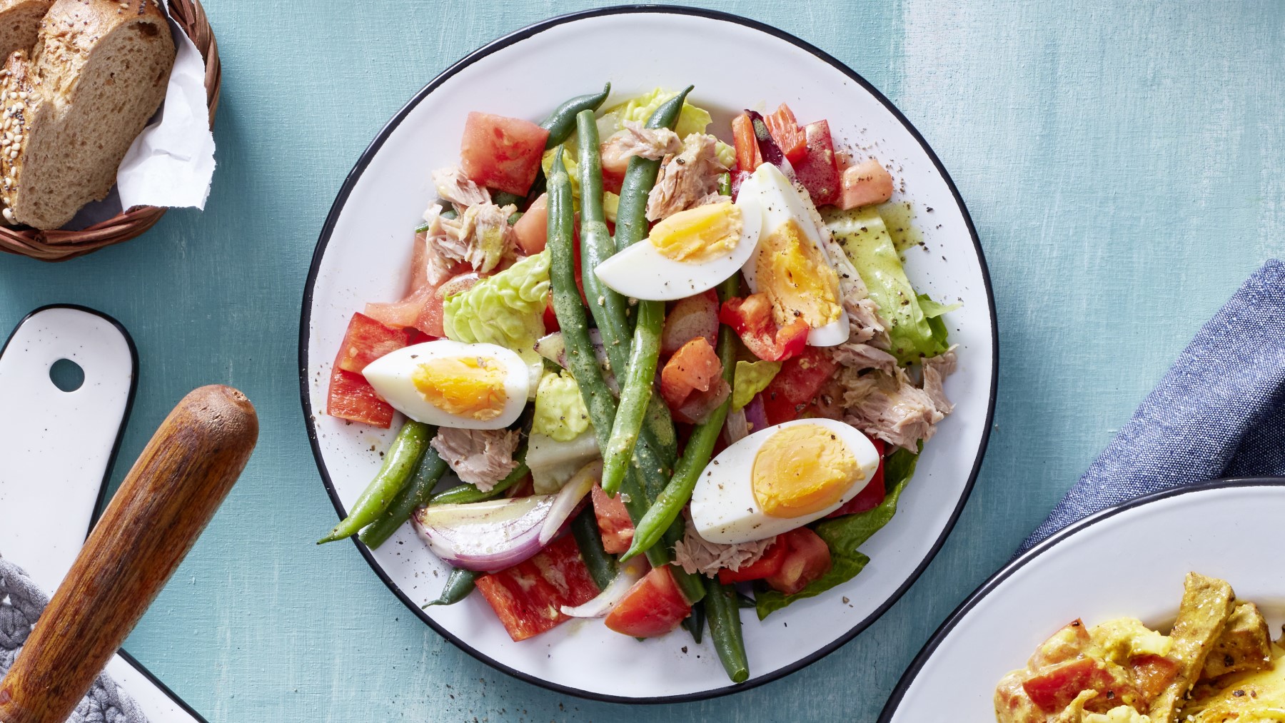 Salat mit Thunfisch und Ei in einer weißen Schüssel auf hellblauem Hintergrund. Links davon etwas Brot.