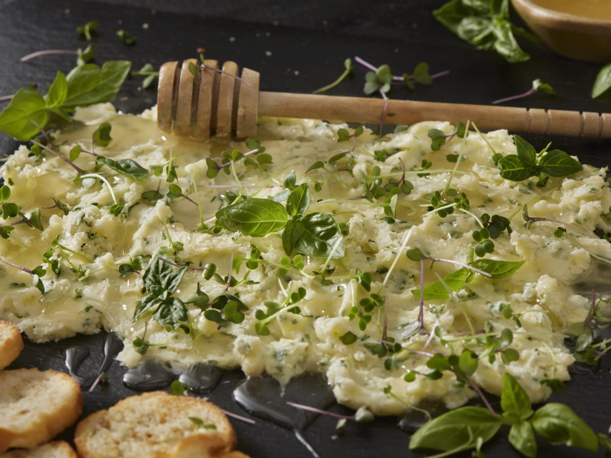 Eine Schiefertafel mit der salzigen Honigbutter und verschiedenen Kräutern und Kresse. Alles von schräg oben auf dunklem Untergrund fotografiert.