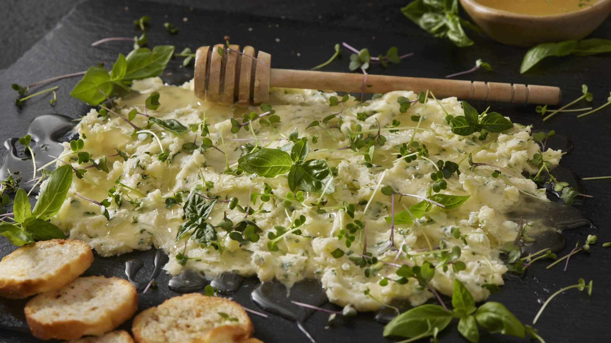 Eine Schiefertafel mit der salzigen Honigbutter und verschiedenen Kräutern und Kresse. Alles von schräg oben auf dunklem Untergrund fotografiert.