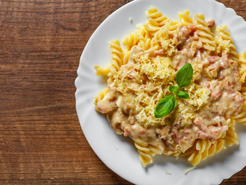 Ein weißer Teller mit Fusilli-Pasta in heller Schinken-Sahne-Sauce, garniert mit geriebenem Käse und einem frischen Basilikumblatt auf Holzuntergrund.