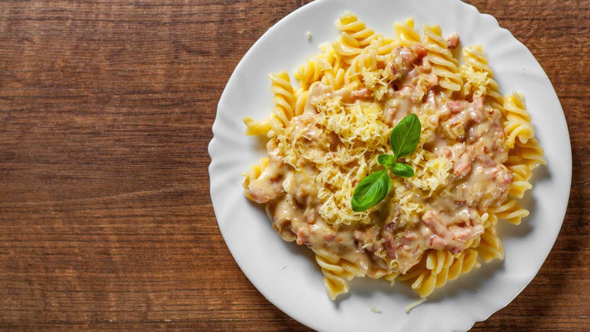 Ein weißer Teller mit Fusilli-Pasta in heller Schinken-Sahne-Sauce, garniert mit geriebenem Käse und einem frischen Basilikumblatt auf Holzuntergrund.