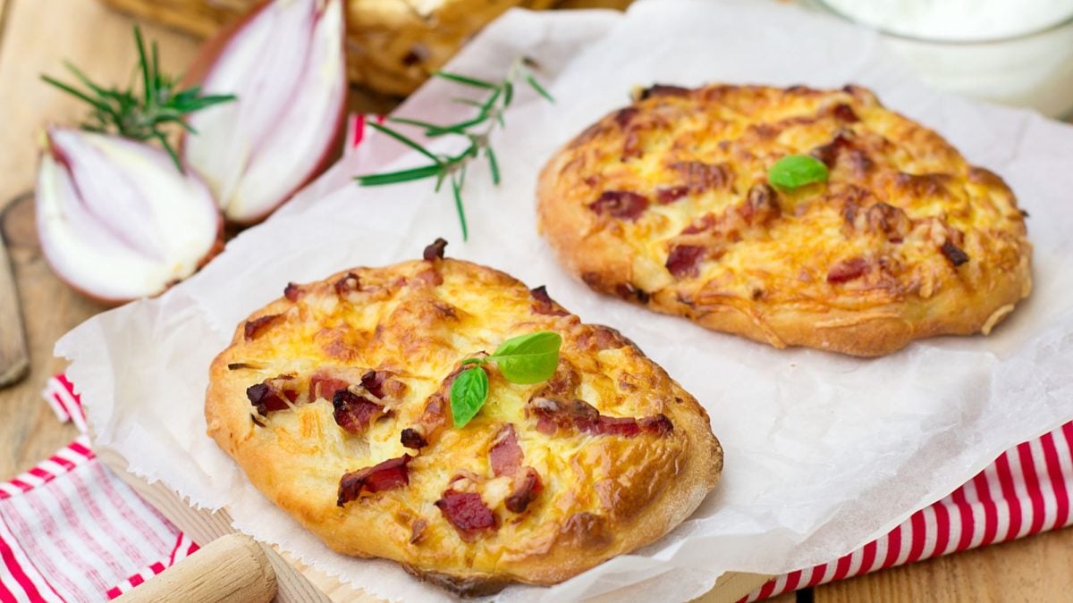 Zwei kleine Pizzabrötchen mit Schmand, Speck und Käse auf Backpapier, dekoriert mit Basilikumblättern, Zwiebeln und Rosmarinzweigen im Hintergrund.