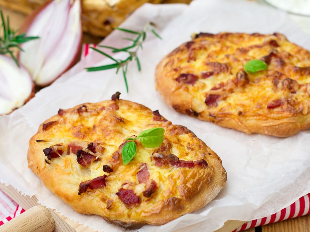 Zwei kleine Pizzabrötchen mit Schmand, Speck und Käse auf Backpapier, dekoriert mit Basilikumblättern, Zwiebeln und Rosmarinzweigen im Hintergrund.