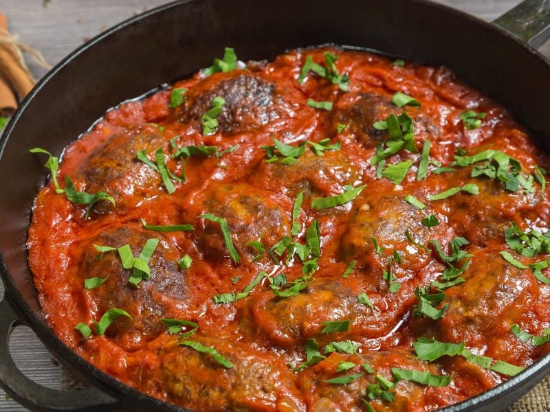 Eine gusseiserne Pfanne mit den Hackbällchen in Tomatensauce von oben auf einem rustikalen Holztisch fotografiert.