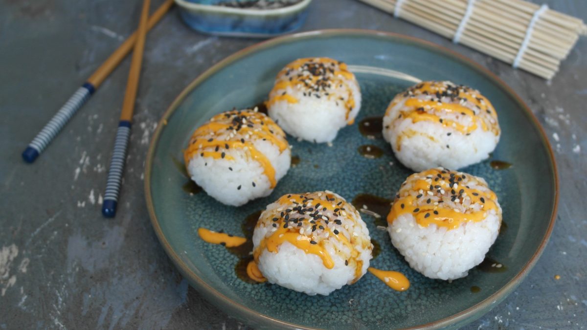 Ein blauer Teller mit den Sushi-Balls auf grauem Untergrund von schräg oben fotografiert. Daneben noch Essstäbchen und Sushi-Matte.