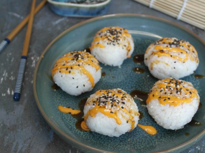 Ein blauer Teller mit den Sushi-Balls auf grauem Untergrund von schräg oben fotografiert. Daneben noch Essstäbchen und Sushi-Matte.