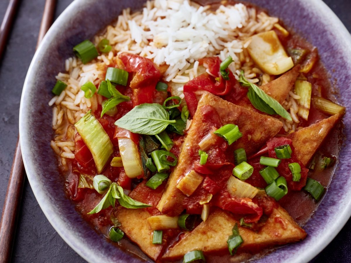 Draufsicht: Tofu süßsauer mit Pak Choi und Paprika auf Reis in einem tiefen Teller, daneben Stäbchen.