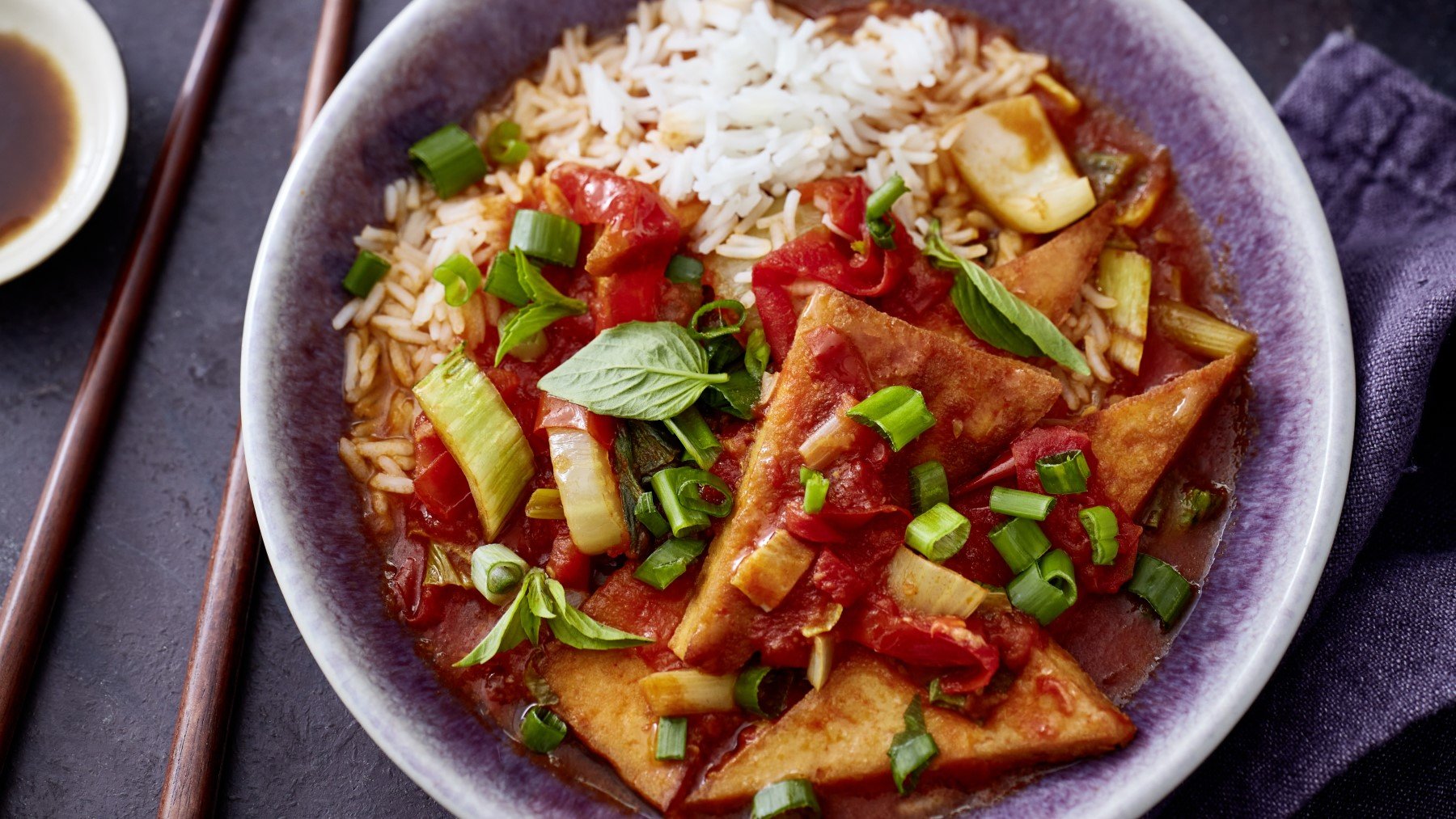 Draufsicht: Tofu süßsauer mit Pak Choi und Paprika auf Reis in einem tiefen Teller, daneben Stäbchen.