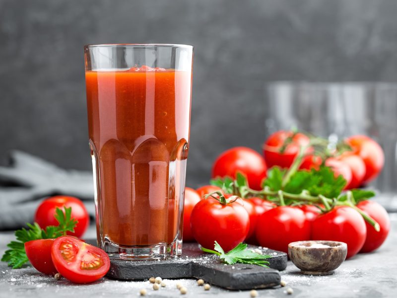 Frisches Glas Tomatensaft neben geschnittenen Tomaten, Kräutern und Gewürzen auf einer rustikalen Steinplatte.