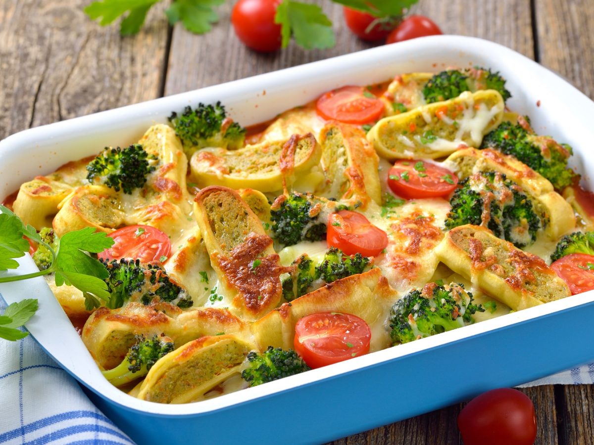 Eine blaue Auflaufform mit dem Maultaschen-Gratin auf einem rustikalen Holztisch mit weiß blauem Geschirrtuch sowie Kirschtomaten und frischer Blattpetersilie.