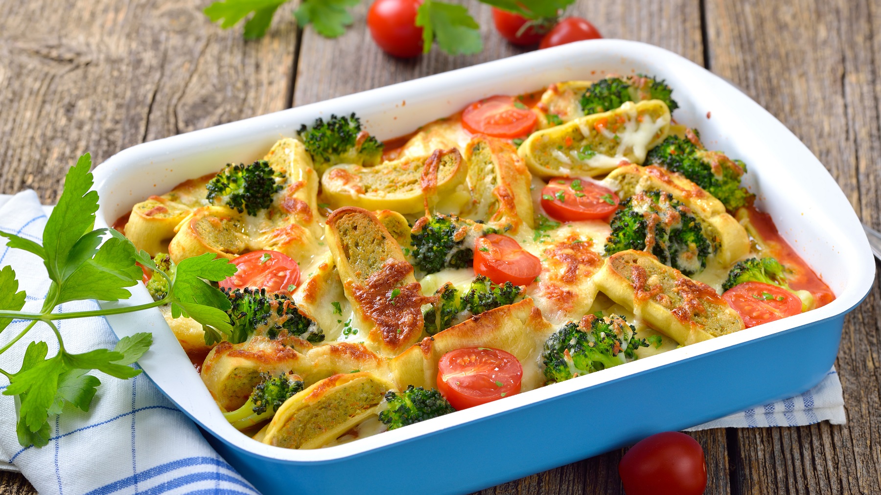 Eine blaue Auflaufform mit dem Maultaschen-Gratin auf einem rustikalen Holztisch mit weiß blauem Geschirrtuch sowie Kirschtomaten und frischer Blattpetersilie.