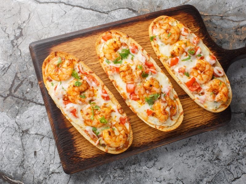 Überbackenes Baguette mit Garnelen auf einem Holzbrett von oben fotografiert.