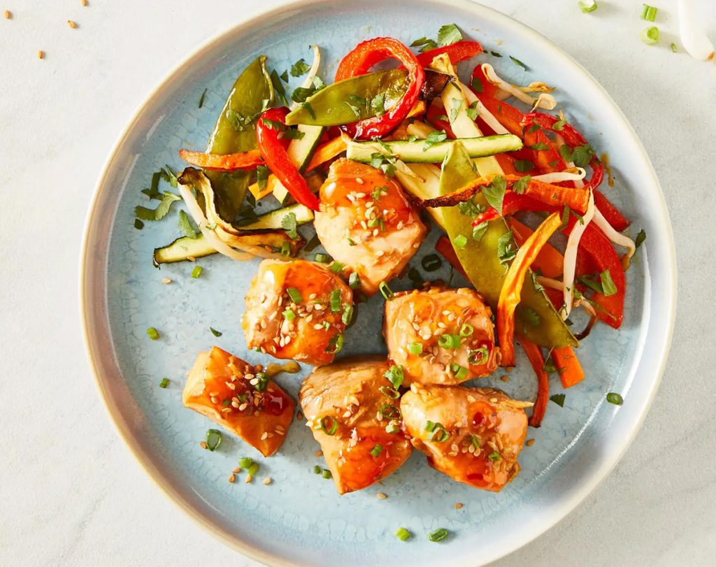 Saftige Lachsstücke mit Sesam auf Gemüsepfanne aus Zuckerschoten, Zucchini und Paprika auf hellblauem Porzellanteller.