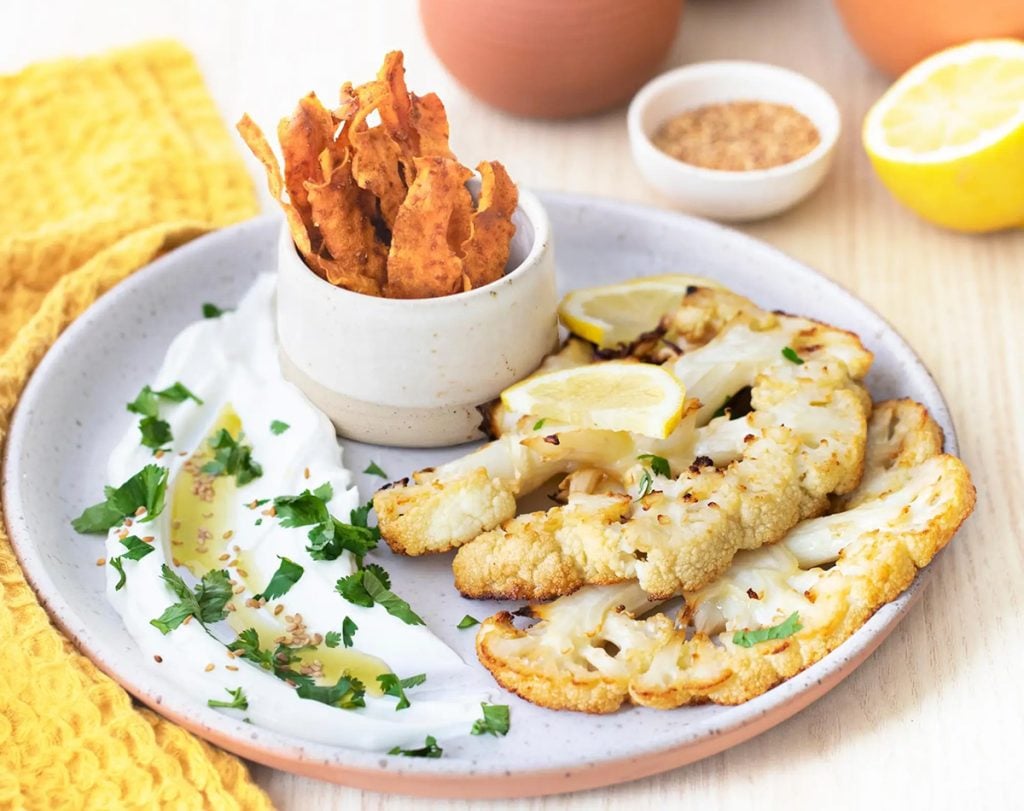 Gegrillte Blumenkohlscheiben mit Zitronenscheiben, serviert auf einem weißen Teller mit Joghurt-Dip, Kräutern und knusprigen Karottenchips.