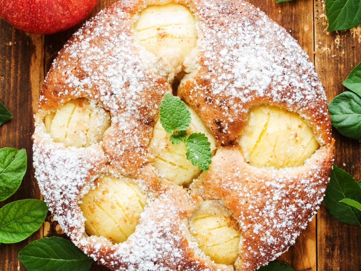 Frisch gebackener Apfelkuchen, garniert mit einem Minzblatt, auf rustikalem Holztisch und grünen Blättern.