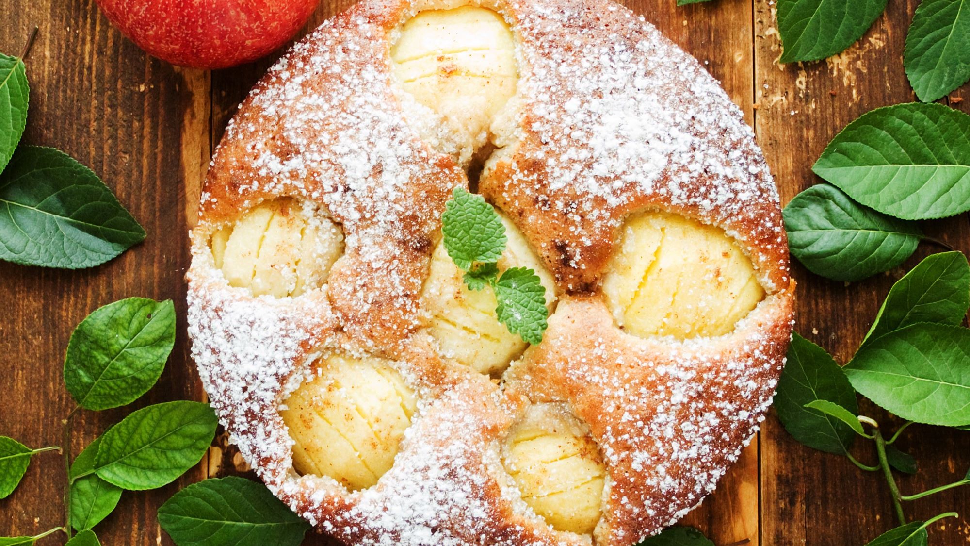 Frisch gebackener Apfelkuchen, garniert mit einem Minzblatt, auf rustikalem Holztisch und grünen Blättern.