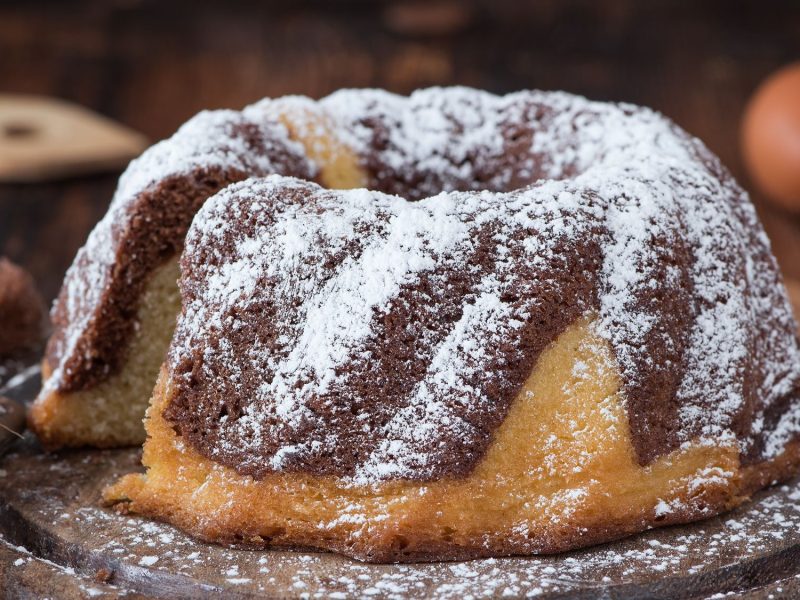 Marmorierter Baileys-Kuchen mit braunen und gelben Streifen, bestäubt mit Puderzucker, auf einem rustikalen Holztisch, daneben zwei Eier und ein Holzlöffel.