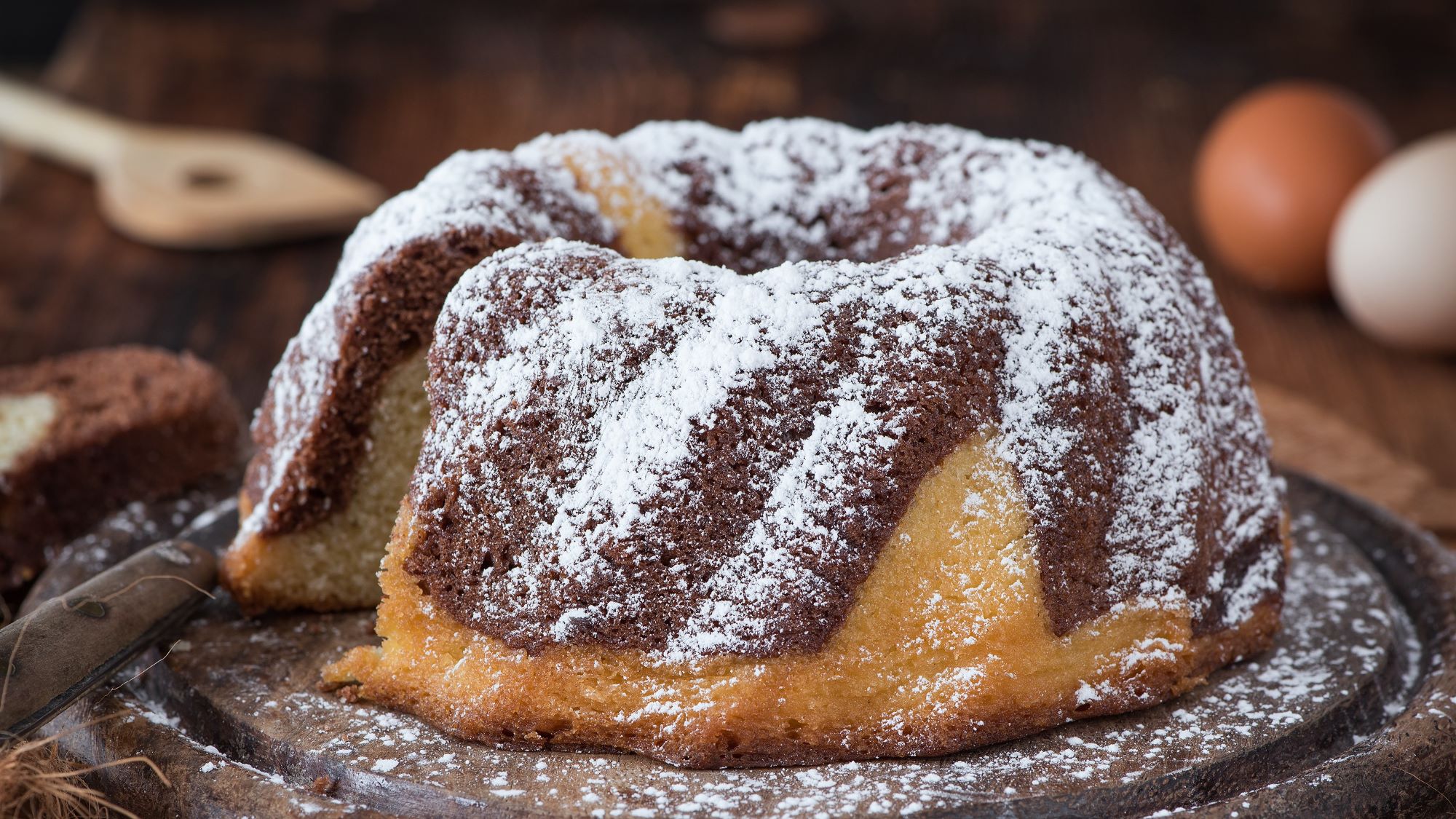 Marmorierter Baileys-Kuchen mit braunen und gelben Streifen, bestäubt mit Puderzucker, auf einem rustikalen Holztisch, daneben zwei Eier und ein Holzlöffel.