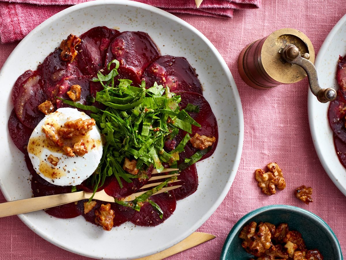 Rote-Bete-Carpaccio mit Walnüssen auf einem weißen Teller auf rosa Hintergrund. Walnüsse, goldenes Besteck und eine goldene Pfeffermühle auf dem Hintergrund verteilt.