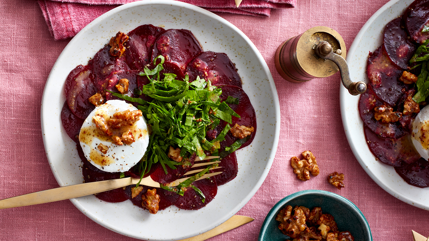 Rote-Bete-Carpaccio mit Walnüssen auf einem weißen Teller auf rosa Hintergrund. Walnüsse, goldenes Besteck und eine goldene Pfeffermühle auf dem Hintergrund verteilt.