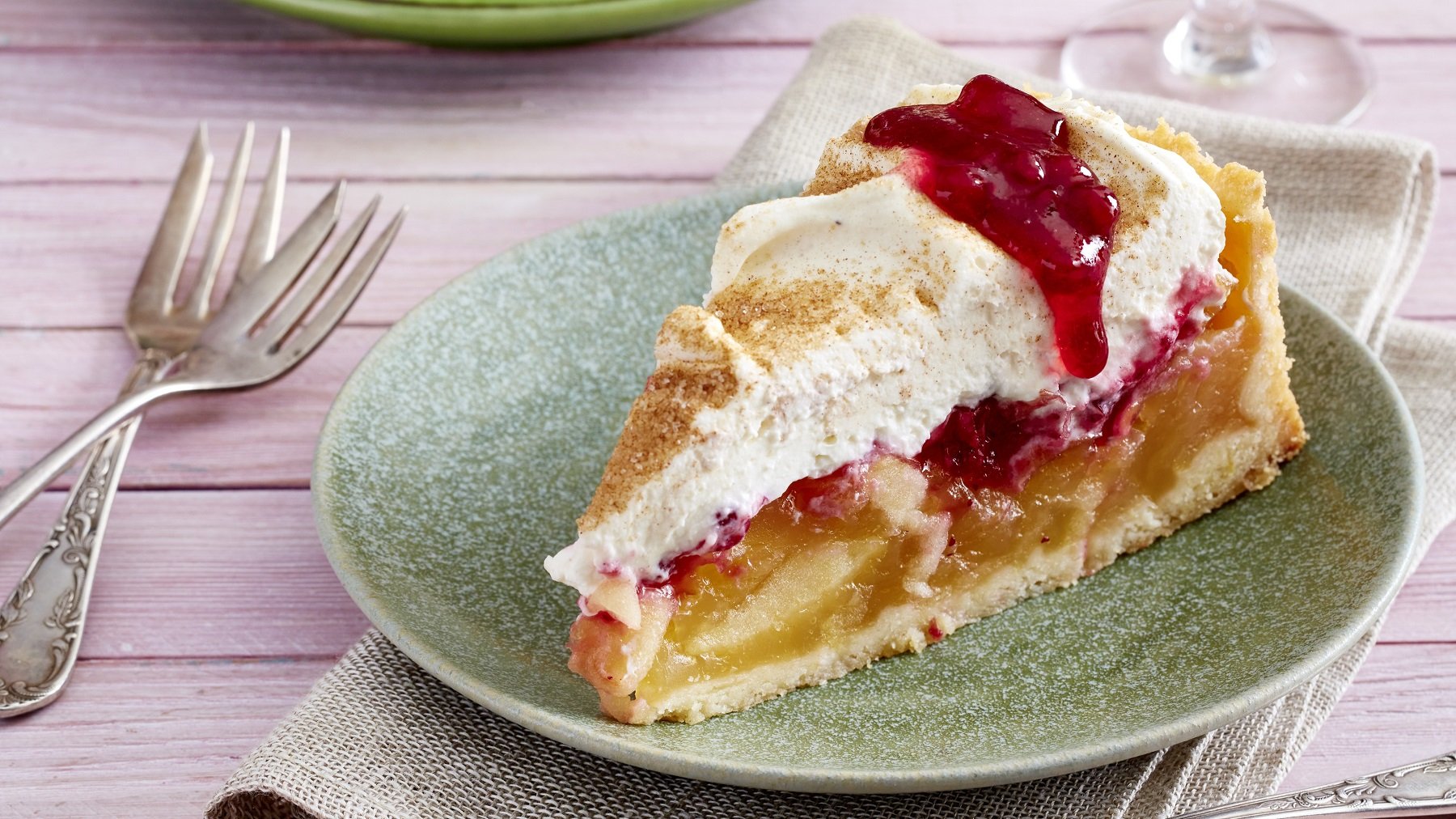 Ein Stück vom Kuchen auf einem grünen Teller und fliederfarbenen Tisch mit zwei kleinen Kuchengabeln. Alles von der Seite fotografiert.