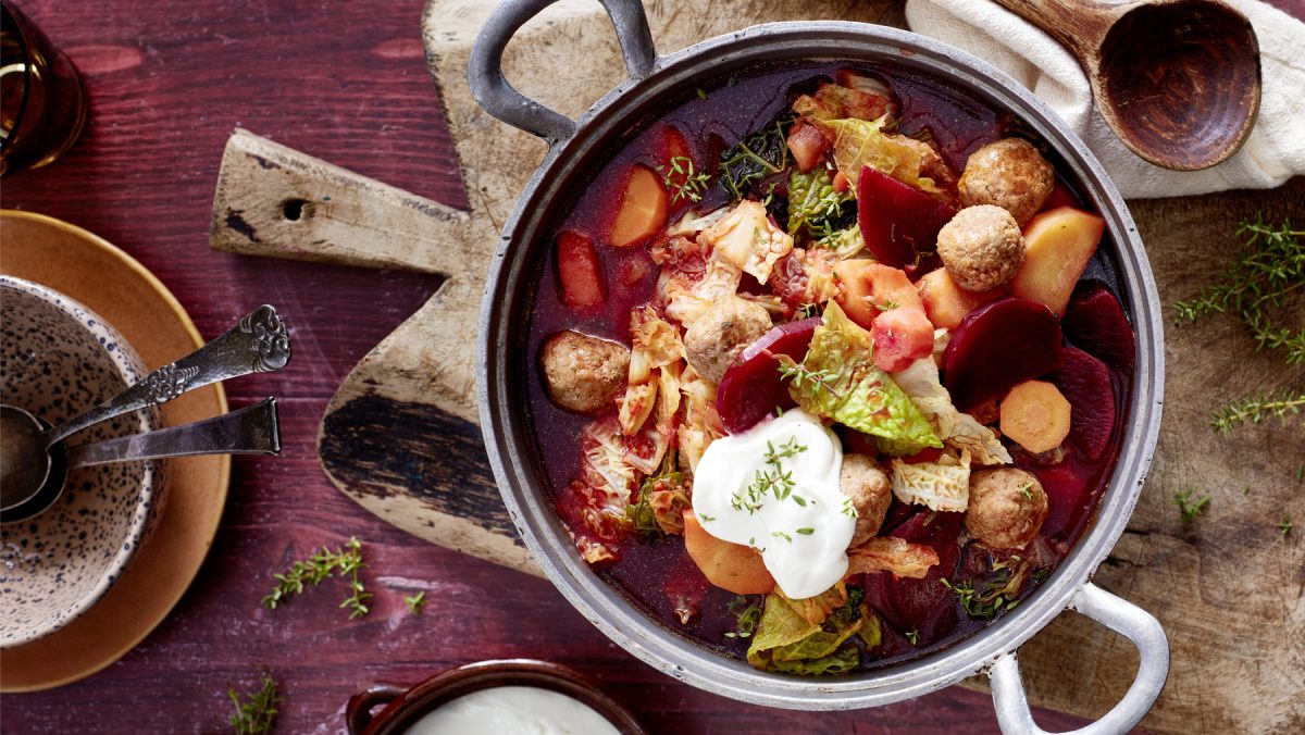 Rote-Bete-Topf mit Hackbällchen auf einem Holzbrett auf dunkelrotem Hintergrund. Links davon eine Schüssel mit Besteck und auf dem Hintergrund Thymian verteilt.