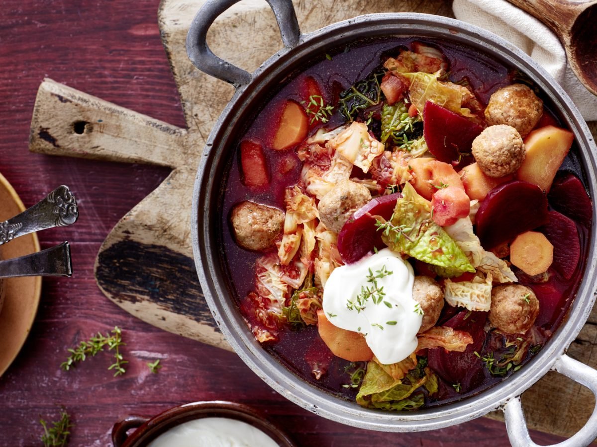 Rote-Bete-Topf mit Hackbällchen auf einem Holzbrett auf dunkelrotem Hintergrund. Links davon eine Schüssel mit Besteck und auf dem Hintergrund Thymian verteilt.