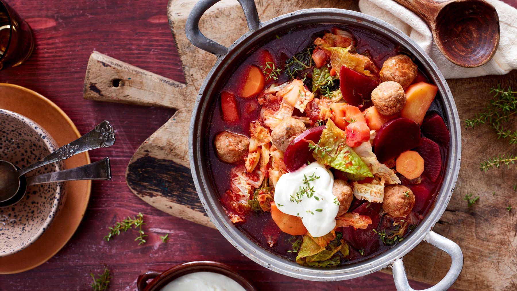 Rote-Bete-Topf mit Hackbällchen auf einem Holzbrett auf dunkelrotem Hintergrund. Links davon eine Schüssel mit Besteck und auf dem Hintergrund Thymian verteilt.