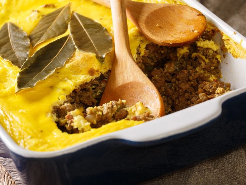 Ein Auflauf mitCurry-Schicht, Hackfleisch und Lorbeerblättern, serviert in einer weißen Auflaufform mit einem Holzlöffel.