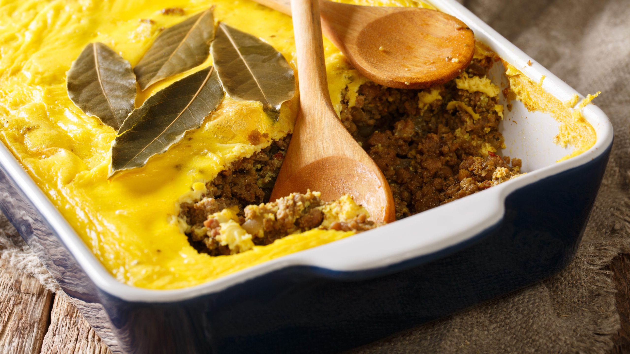 Ein Auflauf mitCurry-Schicht, Hackfleisch und Lorbeerblättern, serviert in einer weißen Auflaufform mit einem Holzlöffel.