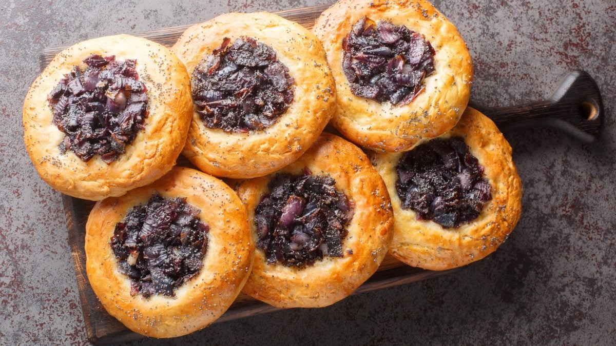 Frische, goldbraune Zwiebelkuchen "Cebularz" mit Mohn auf Holzbrett angerichtet, der Boden zeigt eine rustikale Tischplatte.