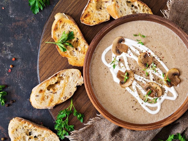 Cremige Champignoncremesuppe mit Sahnedekor, frischen Kräutern und gebratenen Champignons, serviert mit gerösteten Brotscheiben.