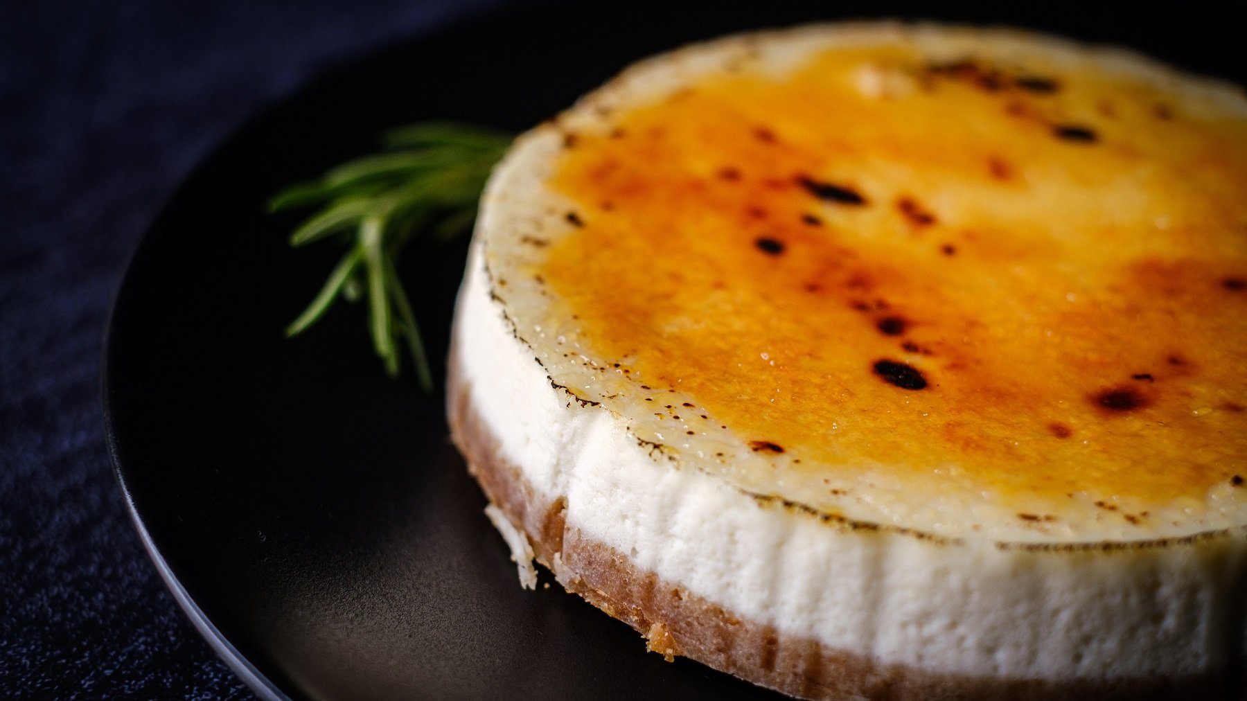 Der Crème brûlée Cheesecake auf einem schwarzen Teller und dunkelblauem Untergrund.