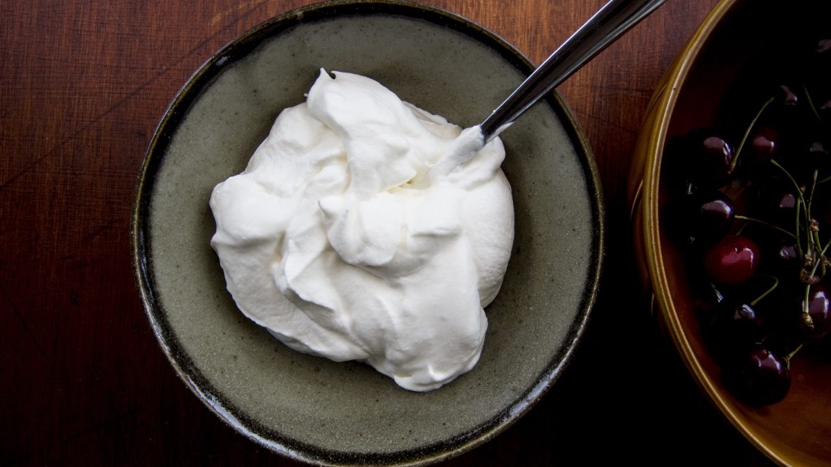 Crème Chantilly in einer grauen Schüssel mit einem Löffel von oben fotografiert, daneben eine Schale mit frischen Kirschen.