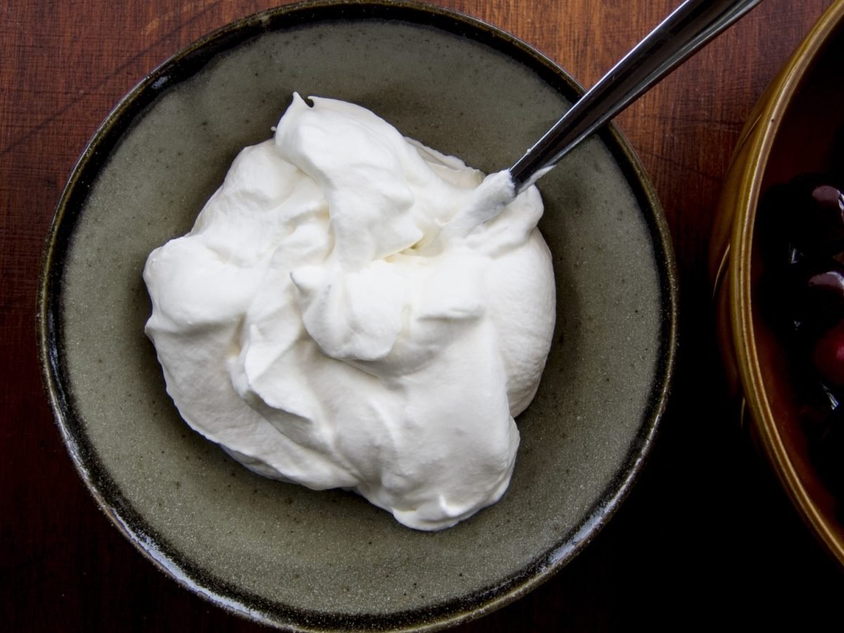 Crème Chantilly in einer grauen Schüssel mit einem Löffel von oben fotografiert, daneben eine Schale mit frischen Kirschen.