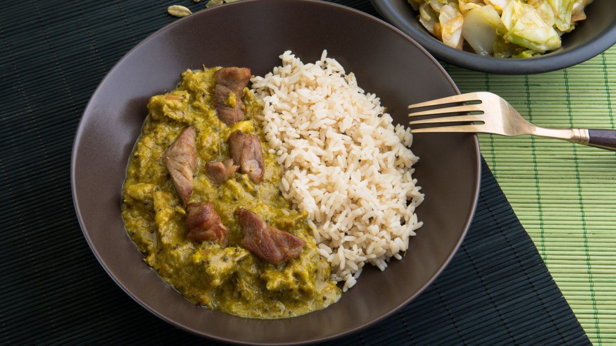 Ein brauner Teller mit Reis undCurry-Geschnetzeltem, angerichtet auf einer schwarzen Bambusmatte mit Gabel.