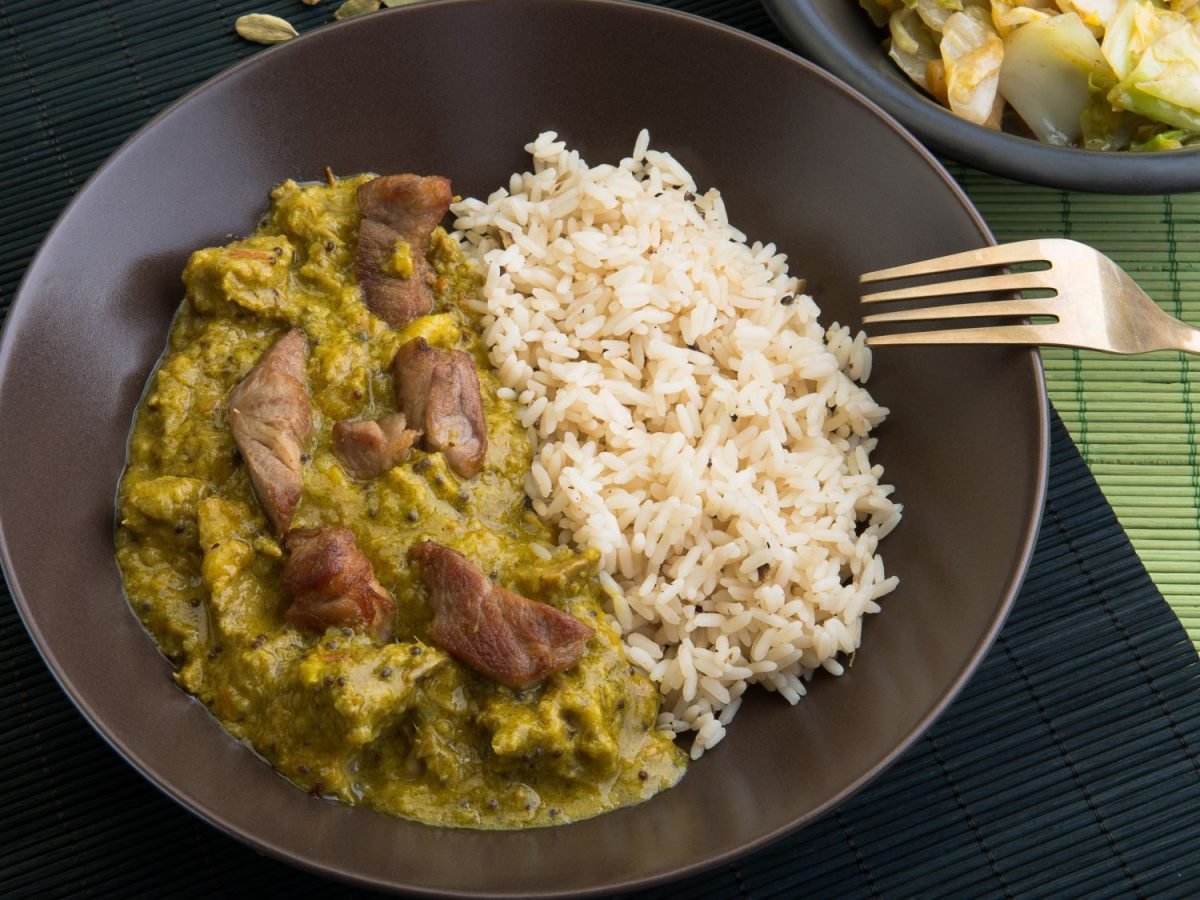 Ein brauner Teller mit Reis undCurry-Geschnetzeltem, angerichtet auf einer schwarzen Bambusmatte mit Gabel.