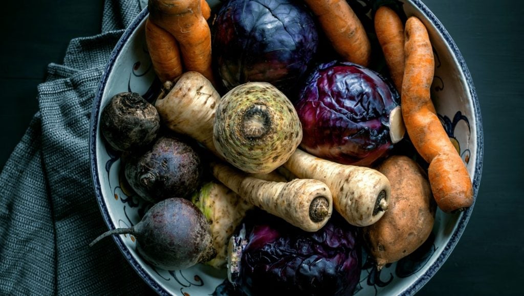 Eine Schale mit Karotten, Pastinaken, Sellerie, Rote Bete und Rotkohl auf einem dunklen Tuch vor schwarzem Hintergrund.