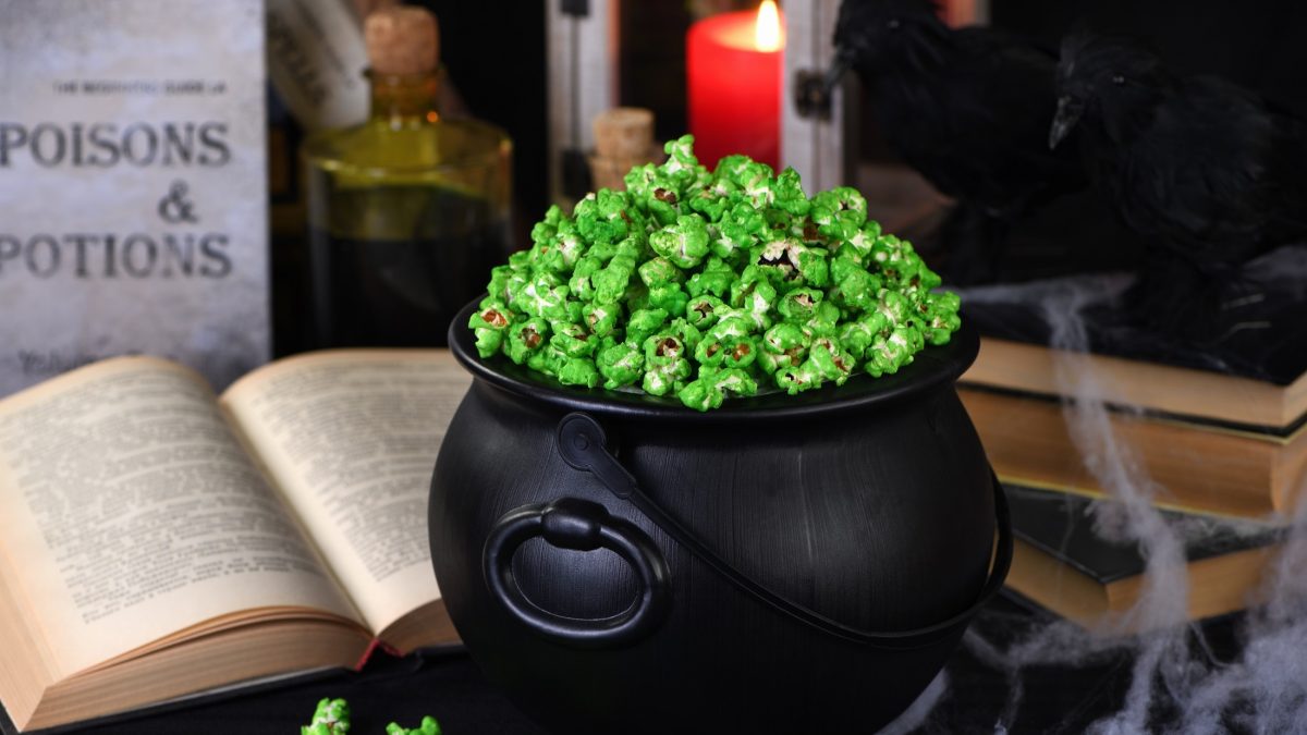 Frontal: Ein rustikaler schwarzer Topf. In dem ist grünes Popcorn, dass an Halloween und am St. Patricks Day selbst gemacht wird. Im Hintergrund sind auf einem Schreibtisch ein dickes altes, aufgeschlagenes Buch und eine brennende Kerze.