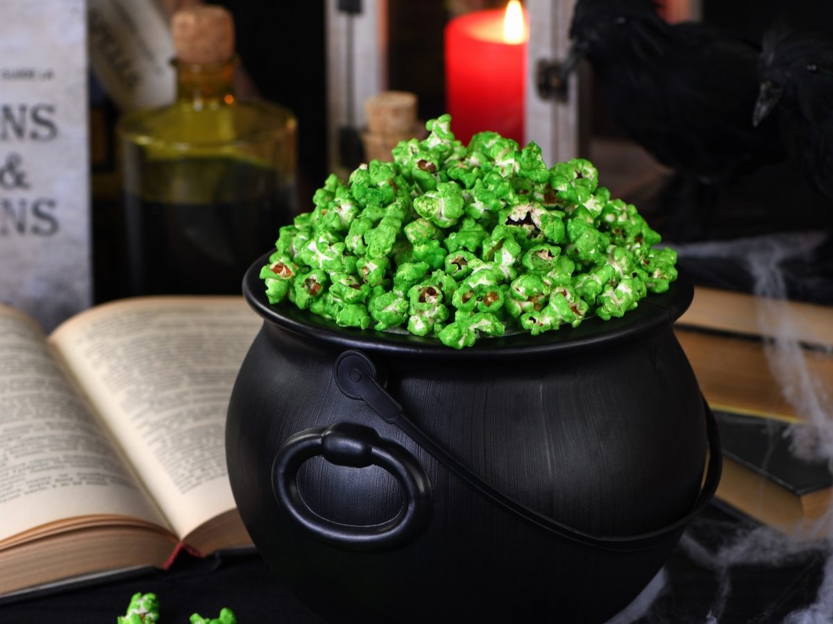 Frontal: Ein rustikaler schwarzer Topf. In dem ist grünes Popcorn, dass an Halloween und am St. Patricks Day selbst gemacht wird. Im Hintergrund sind auf einem Schreibtisch ein dickes altes, aufgeschlagenes Buch und eine brennende Kerze.