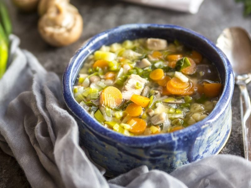 Draufsicht schräg: Ein Topf herbstliche Hühnersuppe auf einem Holztisch. Drum herum liegen frische Zutaten, ein Handtuch und ein Suppenlöffel.